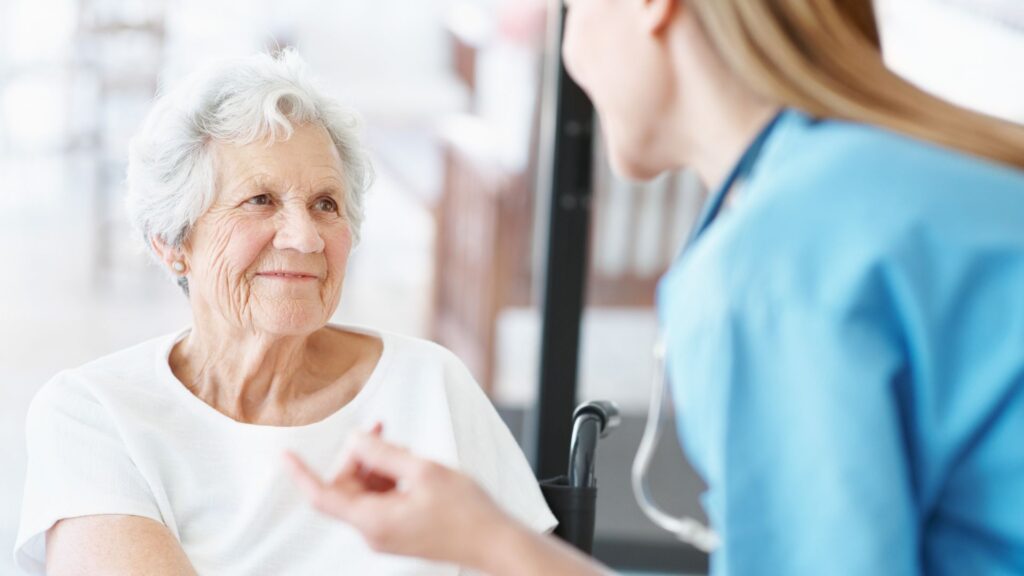 Elderly woman in a white top smiles at a nurse in blue scrubs during a medical check-up in a bright clinic.