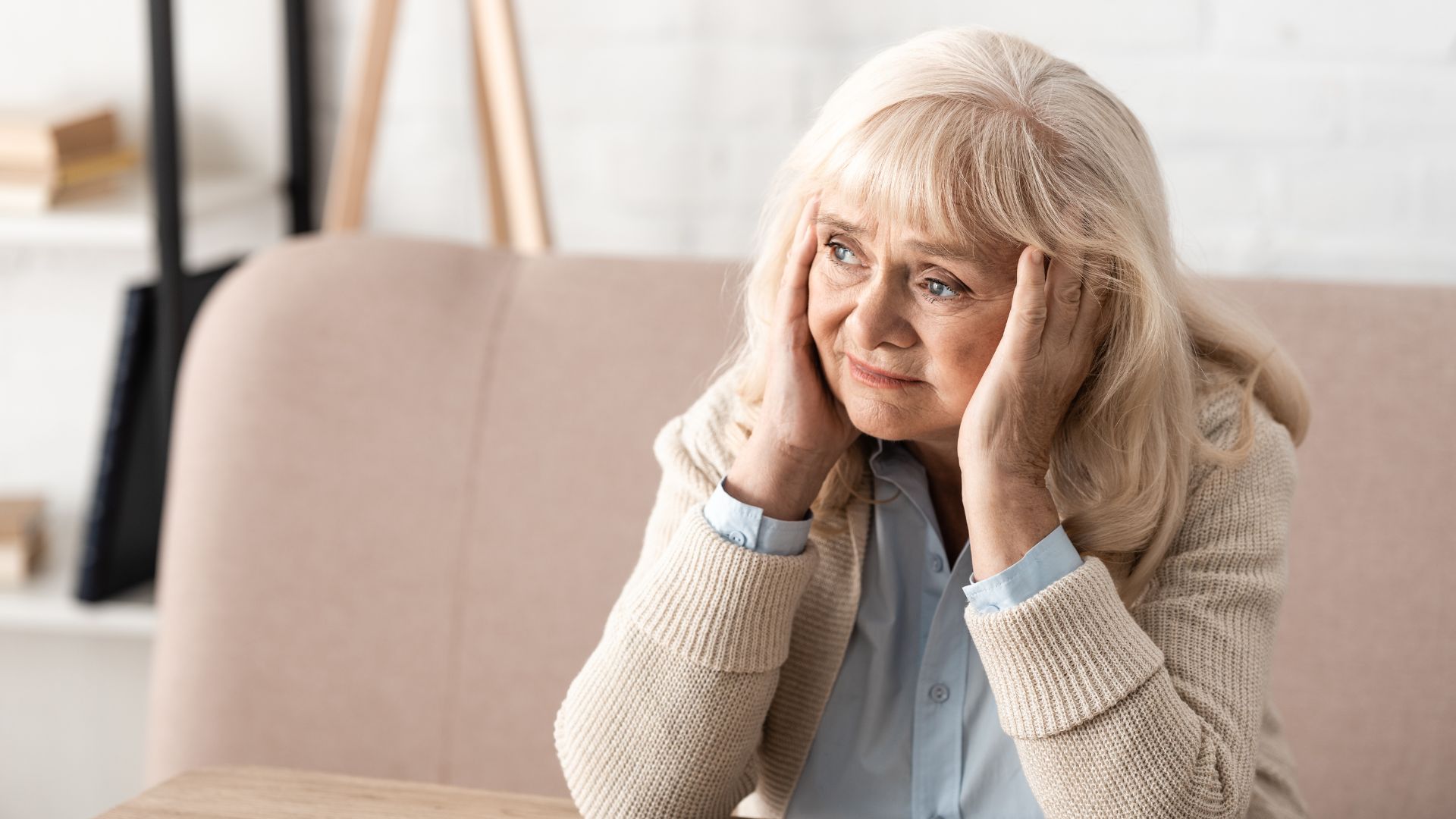 Dementia - RTM Cloud - RTM Cloud Older woman sits at a table, holds her head with both hands, looking worried or stressed.